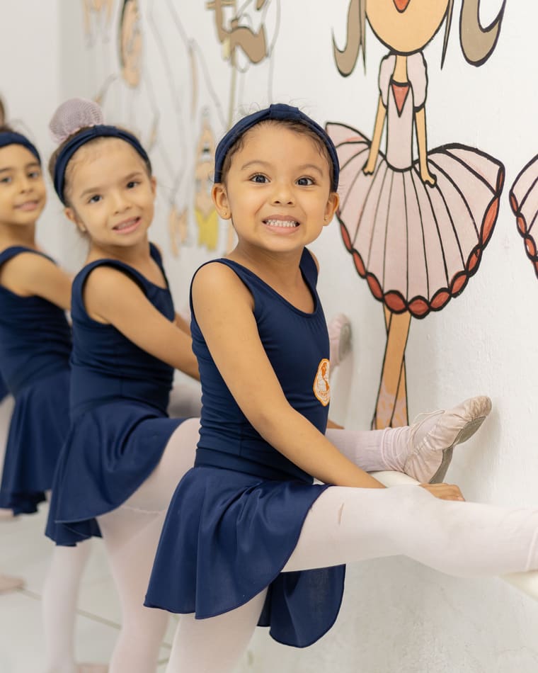 Alunas pequenas de collant azul-marinho alongando na barra da sala de ballet, com bailarinas pintadas na parede