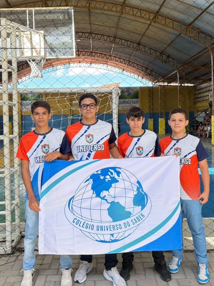 Quatro alunos com o uniforme de jogo segurando a bandeira do Colégio Universo do Saber num ginásio