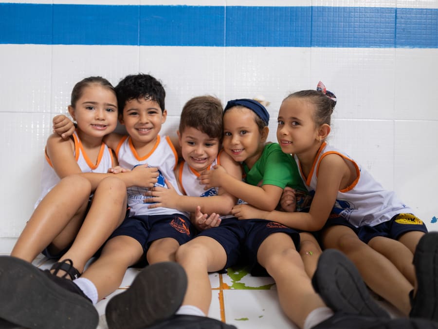 Cinco alunos do Infantil abraçados, sentados no chão e rindo para a foto