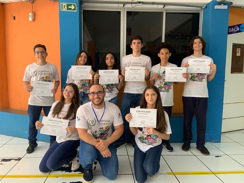 Alunos com a camiseta da Olimpíada de Português segurando os certificados, com o professor agachado à frente