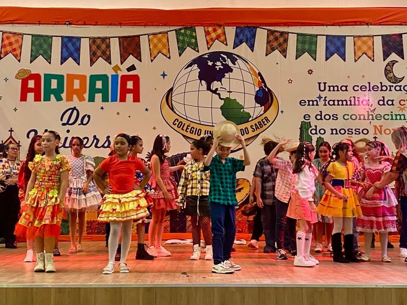 Alunos com roupa de quadrilha dançando no palco do Arraiá do Universo, com as bandeirinhas e o globo do colégio ao fundo