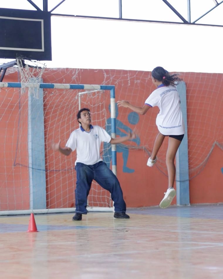 Aluna arremessando no gol de handebol, com o goleiro na frente da parede laranja da quadra