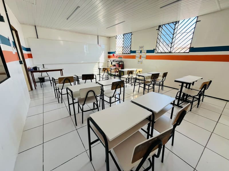 Sala de aula clara, com as carteiras em fileiras e a faixa azul e laranja na parede